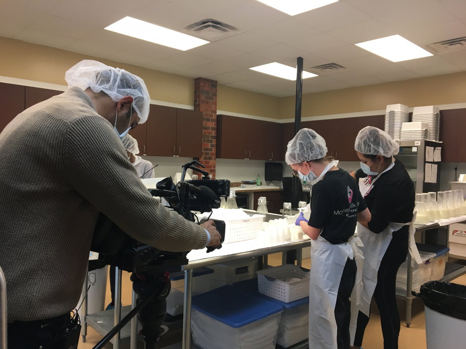 Reporter filming technicians pouring breastmilk into flasks in lab.